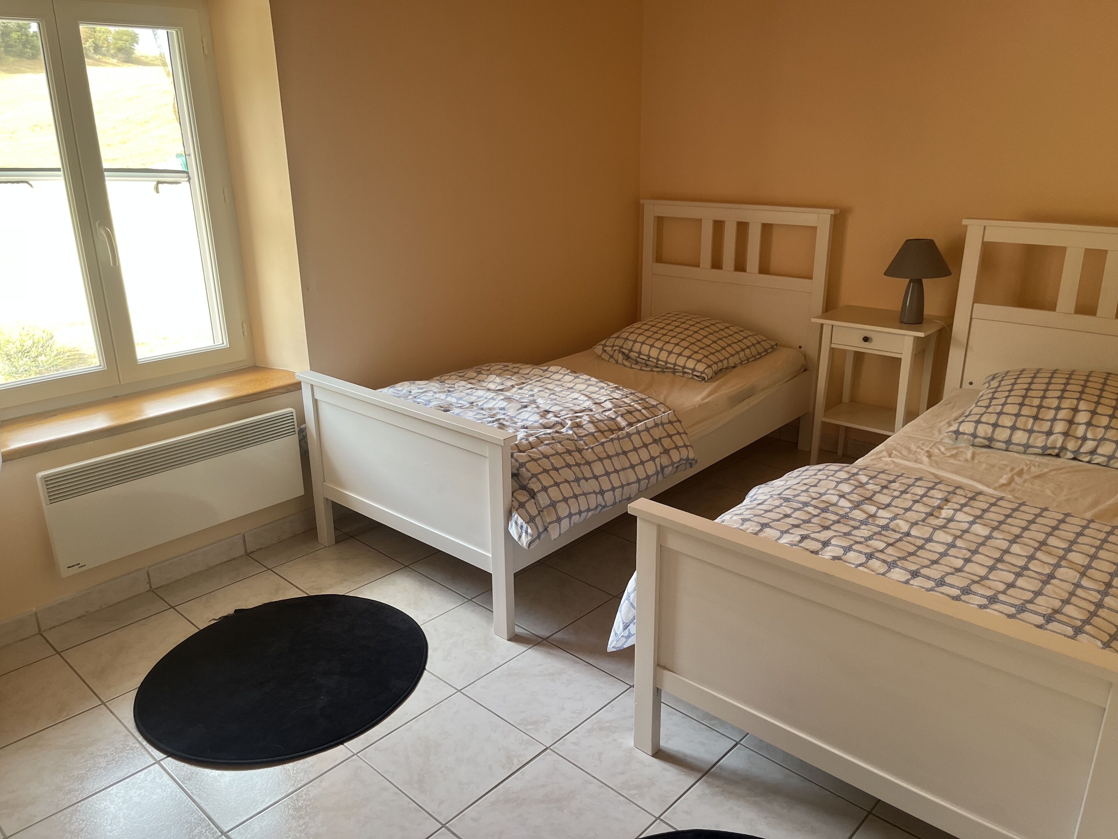 A small, clean room with two single beds, a nightstand with a lamp, and a radiator near the window.