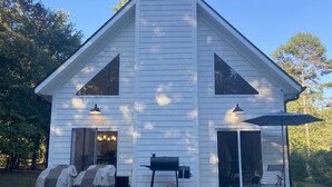 Una casa blanca con una chimenea grande, dos puertas correderas de vidrio, una parrilla cubierta y un patio con sillas.
