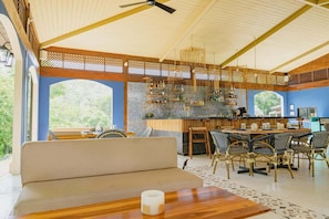 A spacious room with a large window, a ceiling fan, a bar area with shelves, and a seating area with a sofa and chairs.