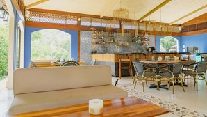 A spacious outdoor dining area with a wooden table and chairs, a bar area with shelves of bottles, and a view of greenery through large windows.