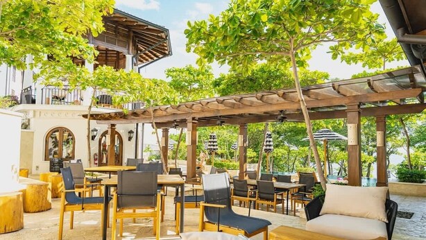 An outdoor patio with wooden tables and chairs, surrounded by greenery and a pergola structure.
