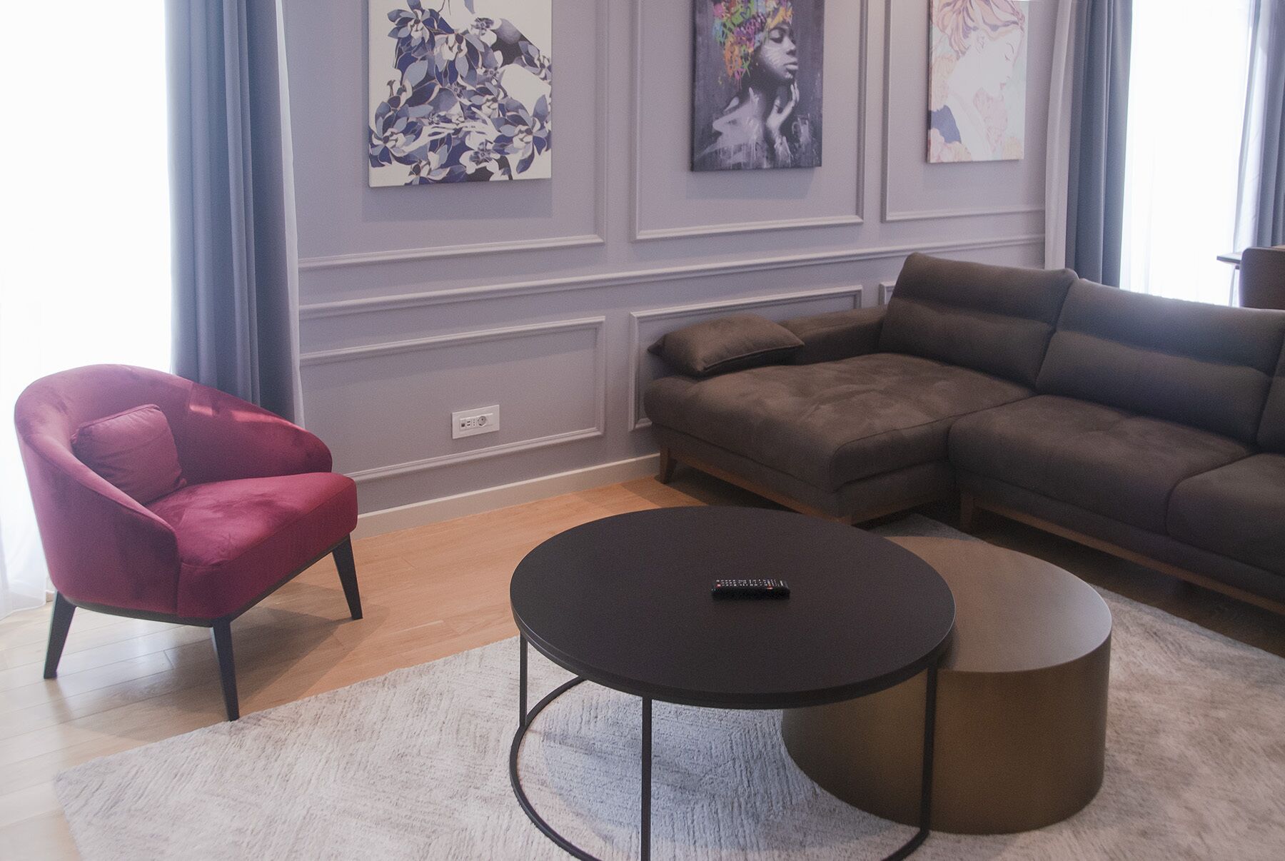 Una sala de estar con un sillón rojo, un sofá seccional marrón, una mesa de centro redonda y una alfombra blanca. Hay tres obras de arte colgadas en la pared sobre el sofá.