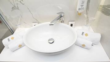 Un lavabo blanc dans la salle de bain, avec un robinet chromé, deux serviettes blanches ornées d’un logo et un flacon de savon liquide fixé au mur.