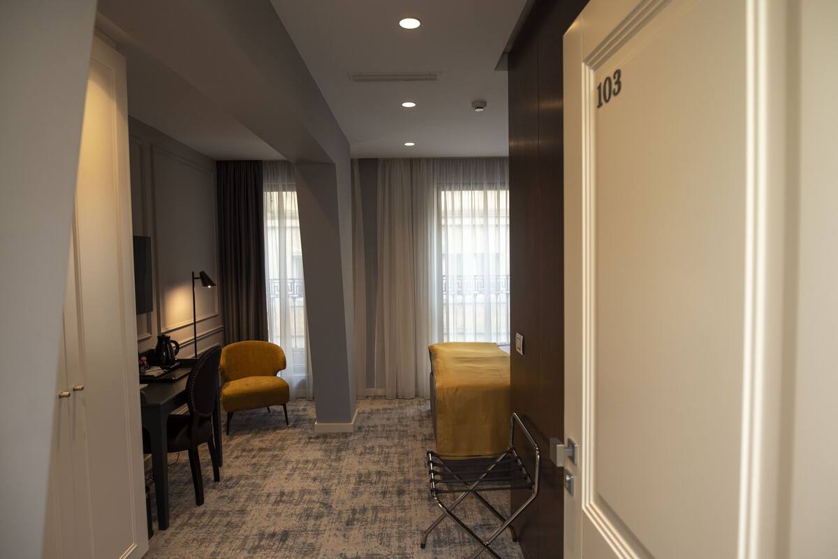 A hotel room with a desk, a yellow armchair, a chair, a coat rack, and a door with the number 103.