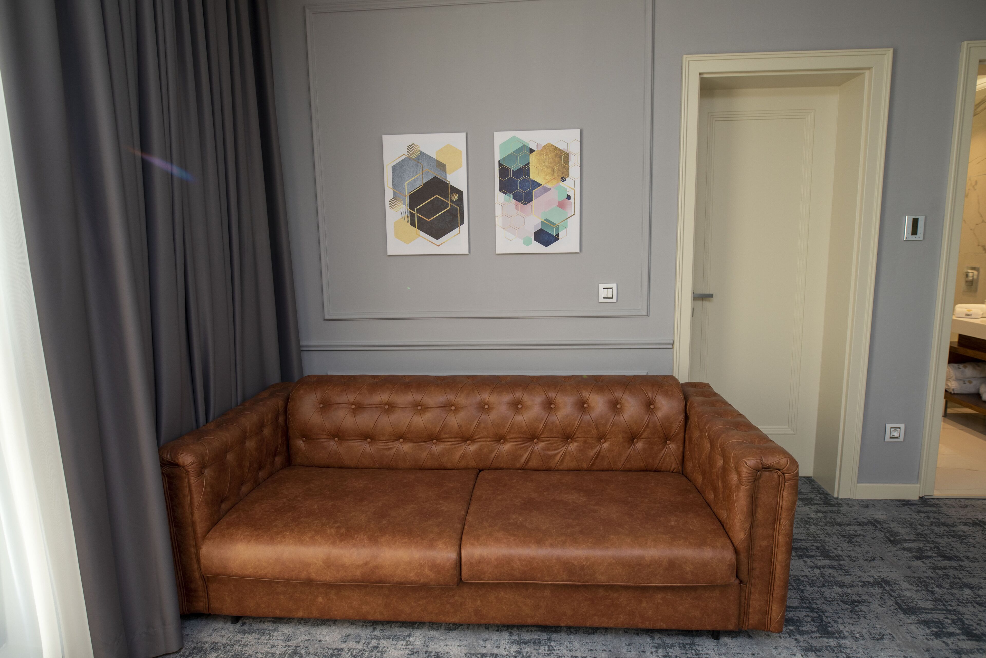 Un sofá de cuero marrón en una habitación con una pared gris y dos cuadros abstractos.