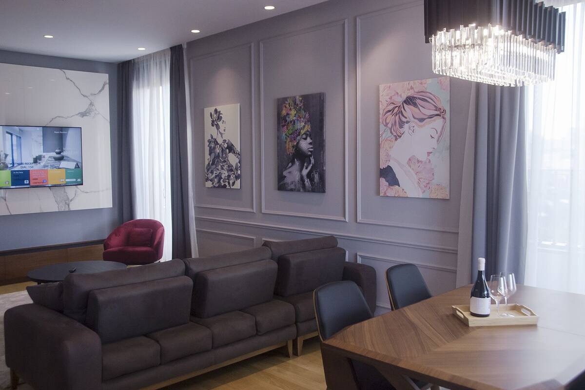 A modern living room with a grey sofa, a wooden dining table with a bottle and glasses, and artwork on the walls.