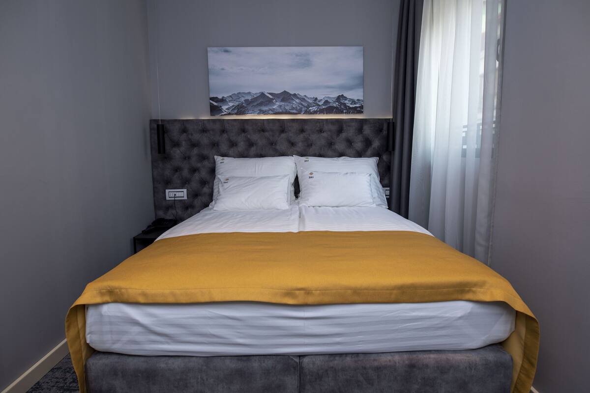 A neatly made bed with a mustard-colored blanket, a tufted headboard, and a framed picture of mountains above it.