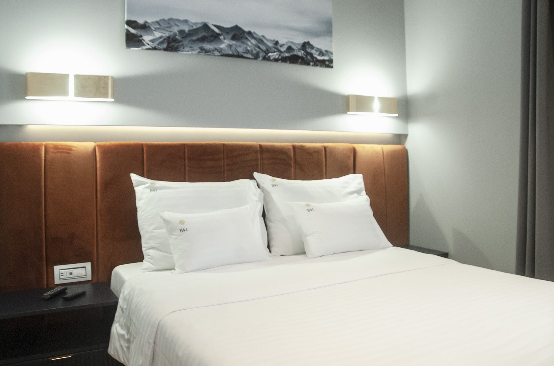 A hotel room with a brown headboard, white bedding, and two wall-mounted lights.