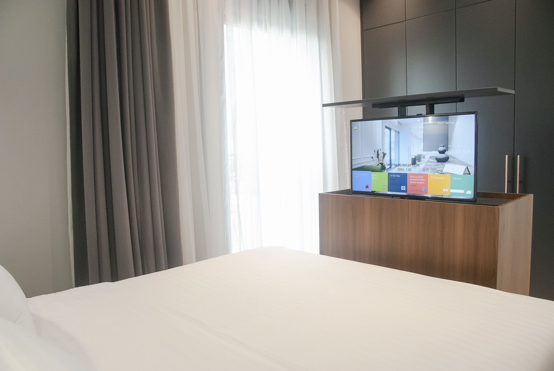 Una habitación de hotel con cama, televisión montada en un mueble de madera y ventana con cortinas.