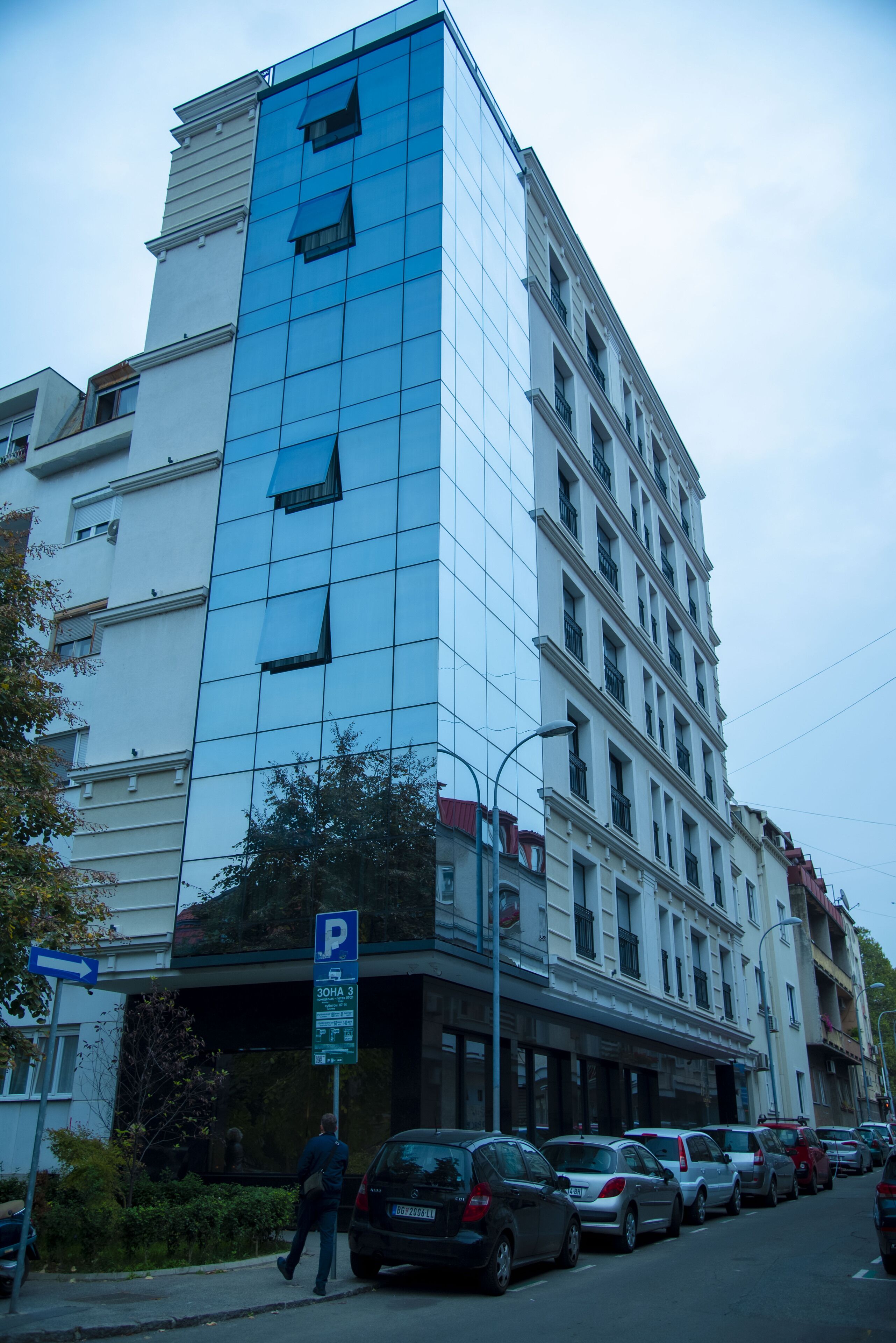 A multi-story building with a modern glass facade, a parking sign, and several parked cars along the street.