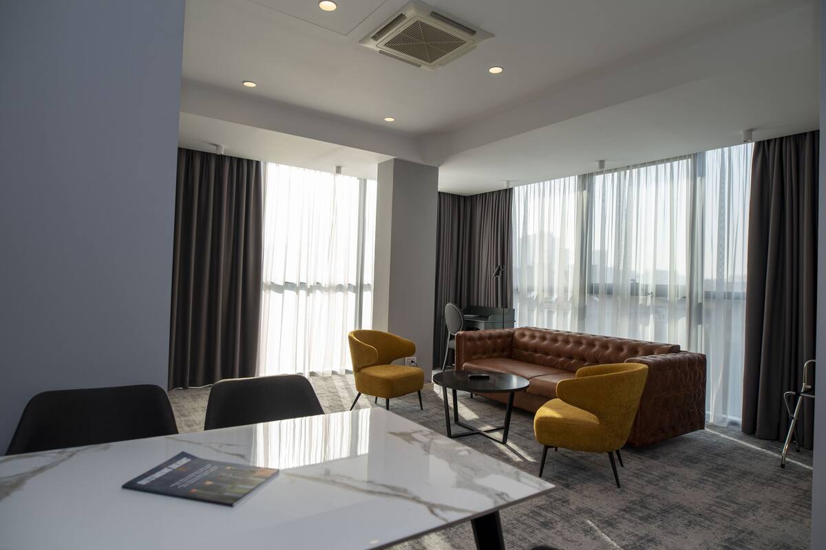 A modern hotel room with a large window, a sofa, two armchairs, a small table, and a mirror.