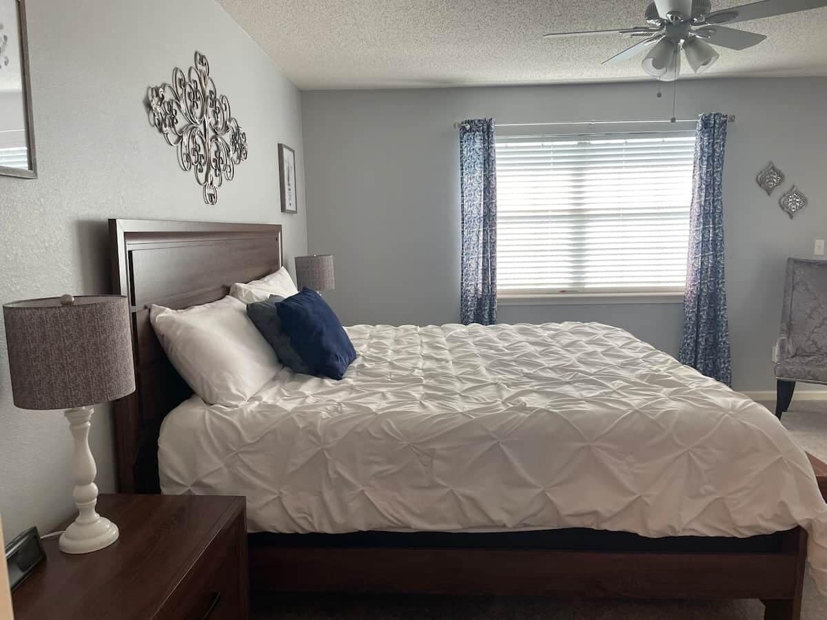 Une chambre à coucher avec un lit, une table de chevet, une lampe, un ventilateur de plafond et une fenêtre avec des rideaux.