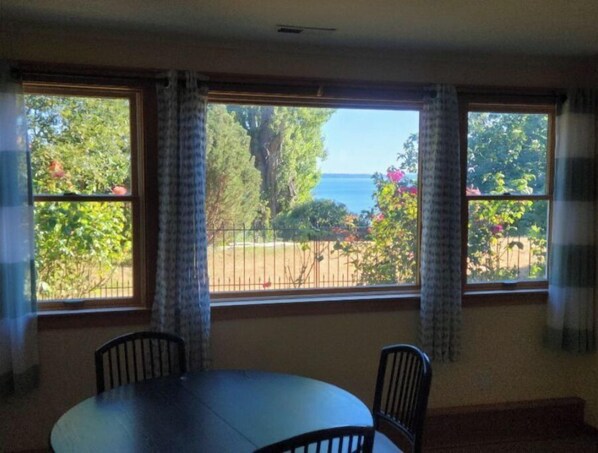 Un comedor con una mesa y sillas, ventanas amplias con cortinas, y vistas a un jardín y al mar.
