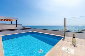 A modern outdoor pool with a clear view of the sea, a wooden deck, and a bottle of champagne.