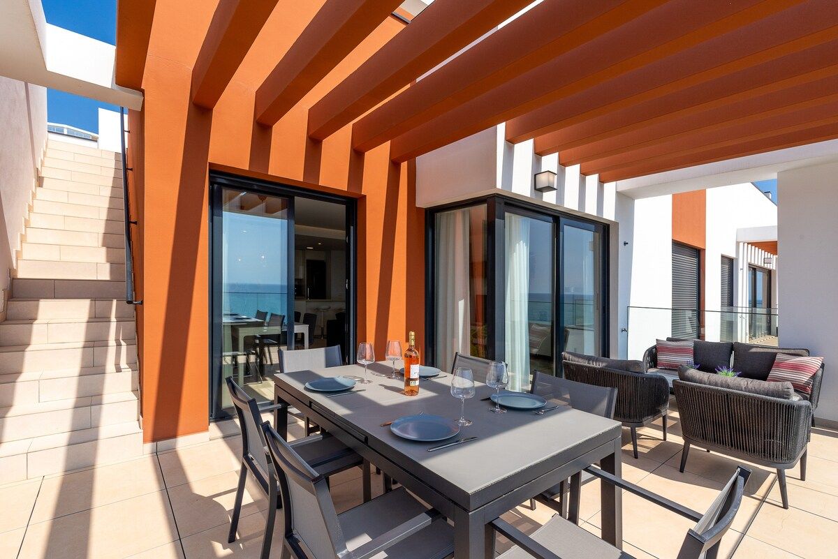 An outdoor dining area with a table set for two, a sofa, and a view of the sea.