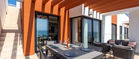 An outdoor dining area with a table set for two, a sofa, and a view of the sea.