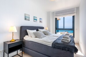 A modern bedroom with a large bed, bedside lamps, a view of the sea, and a window with a view of buildings.