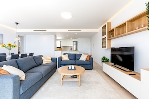 Un salon moderne avec un canapé, une table basse, un téléviseur et une cuisine en arrière-plan.