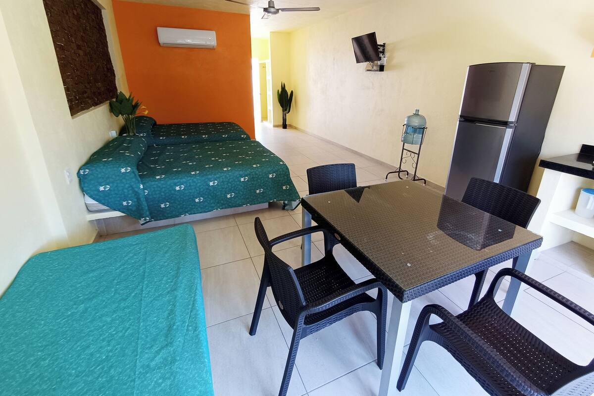 A compact room with a bed, a table, chairs, a refrigerator, a water dispenser, and a ceiling fan.