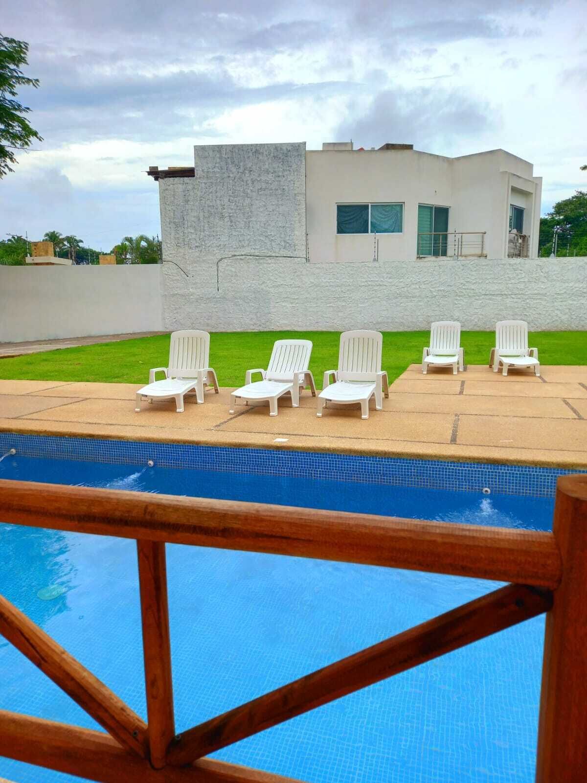 Una piscina con seis sillas de jardín blancas dispuestas alrededor, un edificio moderno con grandes ventanales al fondo y un césped.