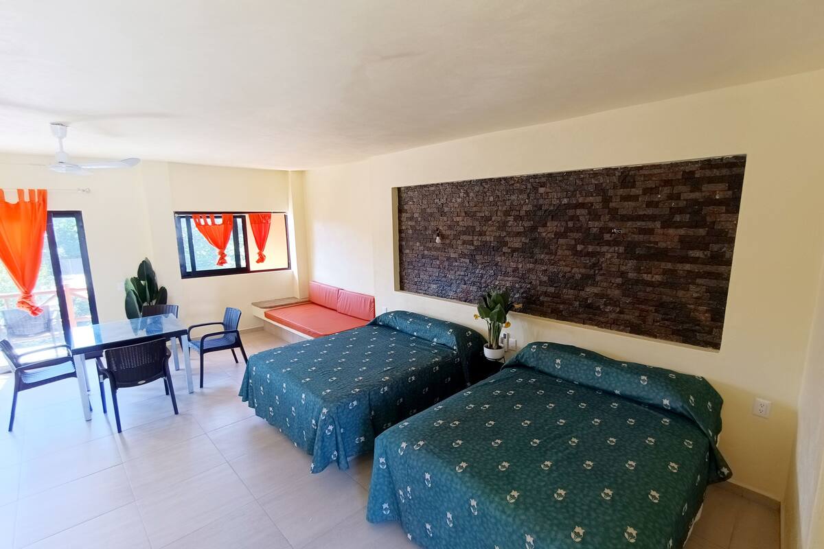 A room with two beds, a desk, chairs, a window with curtains, and a brick accent wall.