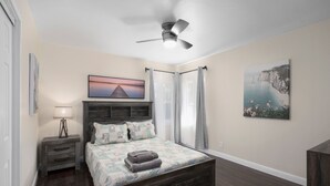 Une chambre à coucher comprenant un lit, une table de chevet, une lampe, un ventilateur de plafond et des œuvres d’art accrochées aux murs.