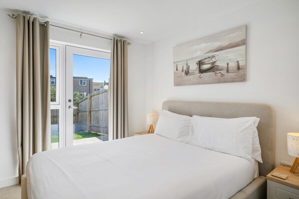 A bedroom with a bed, a nightstand, a lamp, a painting, and a view of the outside.