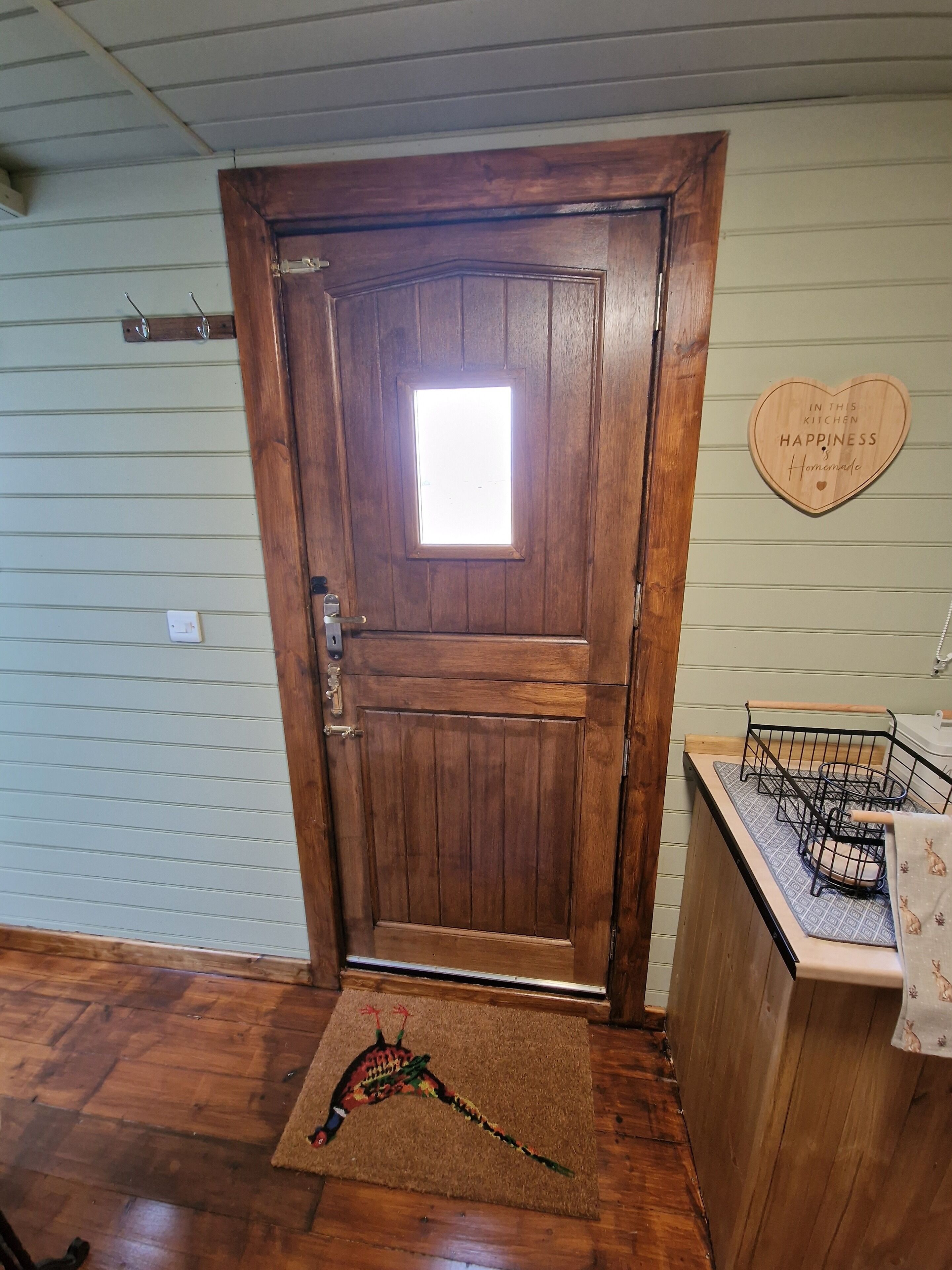 A wooden door with a small window, a heart-shaped sign on the wall, and a doormat with a colorful design.