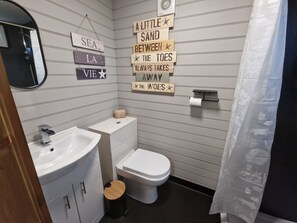 Een badkamer met een wastafel, toilet en een spiegel. Er hangen houten bordjes aan de muur met teksten zoals 'EEN LITTEL ZAND TUSSEN DE TEENEN VERWIJDERD DE TEENEN'.