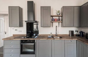Una cocina moderna con armarios grises, horno integrado, cafetera y una estantería con libros y una planta.