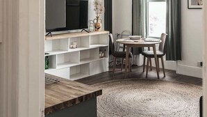 Een moderne woonkamer met een televisie, houten planken, een eettafel en een groot geweven vloerkleed.
