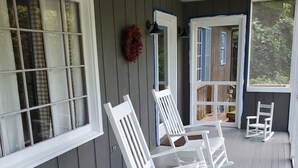 A porch with white rocking chairs, a wreath, and a view of a wooden house.