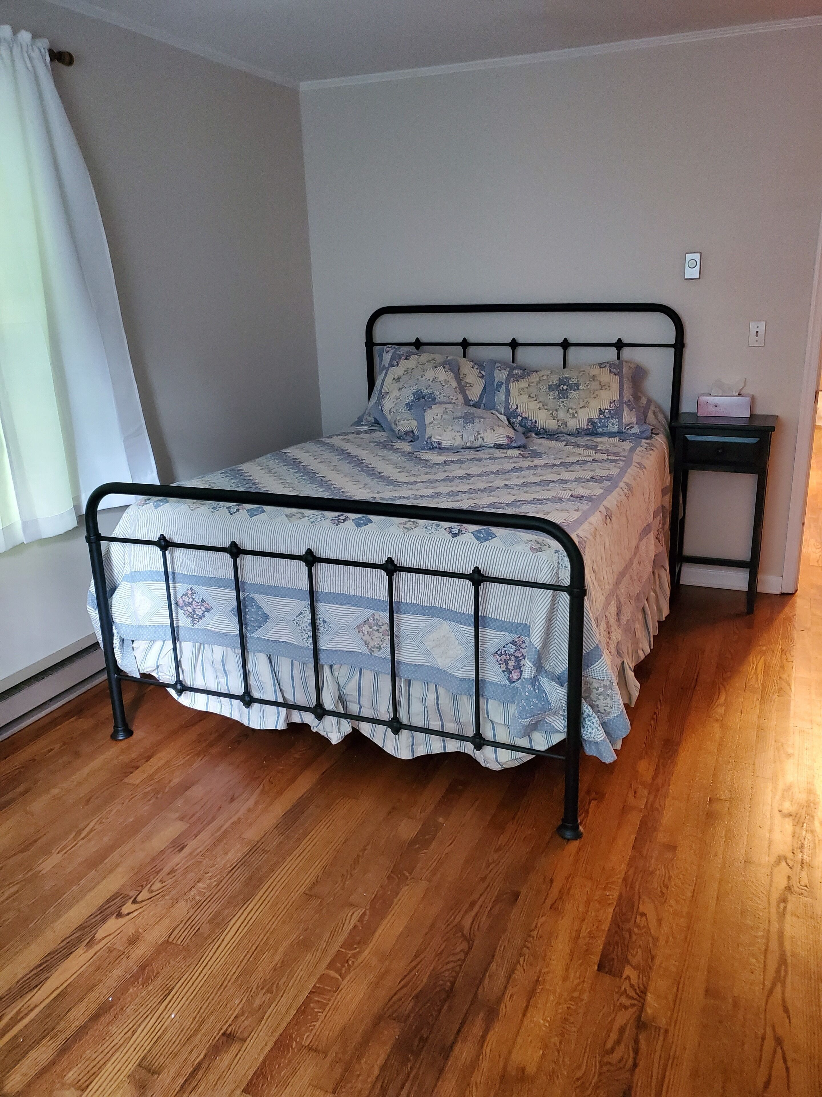 A single bed with a metal frame and a patterned bedspread, a bedside table with a tissue box, and a wooden floor.