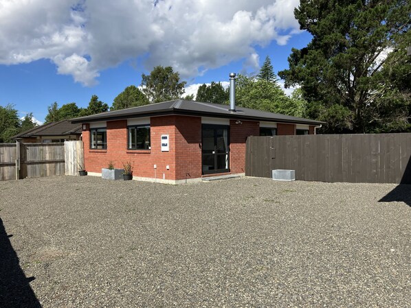 A small brick building with a metal roof, a wooden fence, and a gravel driveway.