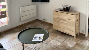 Ett vardagsrum med en platt-TV som är monterad på väggen, en byrå i trä, ett bord med glasskiva och en broschyr på, samt ett element.