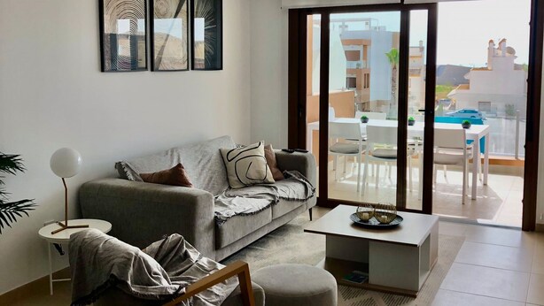 A modern living room with a grey sofa, a coffee table, and a side table with a lamp. There is a large window with a view of the outside.