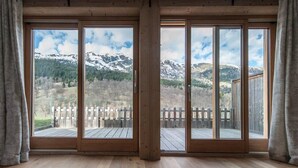 Ein Zimmer mit großen Schiebeglasfenstern, die auf eine Terrasse mit Holzwertung führen und einen Blick auf schneebedeckte Berge und eine bewaldete Landschaft bieten.