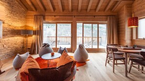 Ein gemütliches Zimmer mit Holzboden, Steinwand, großen Fenstern, einem Esstisch, Stühlen und Sitzsäcken.