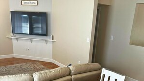 Una sala de estar con un sofá seccional color beige, una mesa de comedor de madera con sillas, un televisor montado en la pared y un letrero que dice "Beach House".