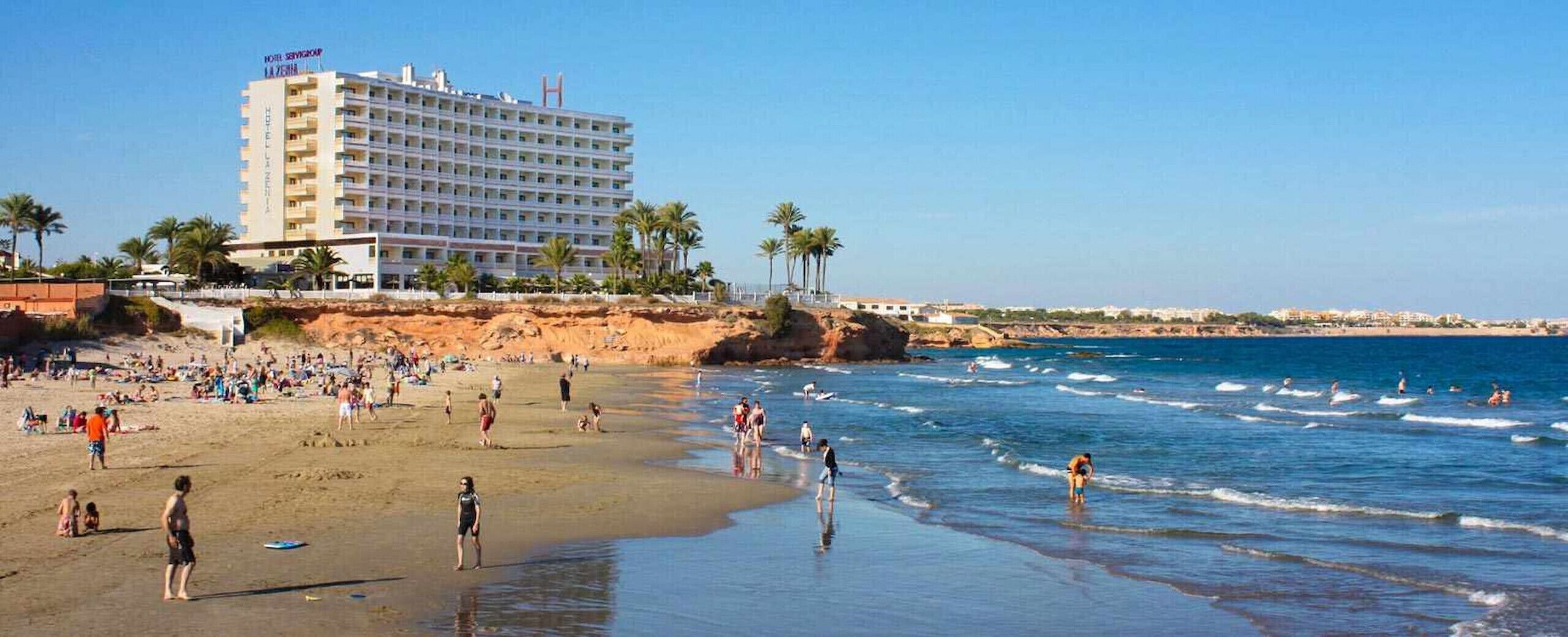 A hotel with multiple floors, a beach in the foreground, and palm trees.