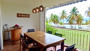 A dining area with a wooden table and chairs, a small bar area, and a view of the ocean with palm trees.