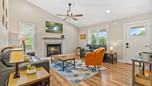A modern living room with a fireplace, a ceiling fan, and a view of the outdoors through the windows. - New home w/hot tub, fireplace, firepit - Less than 1/2 mi to Main St. (Waynesville)