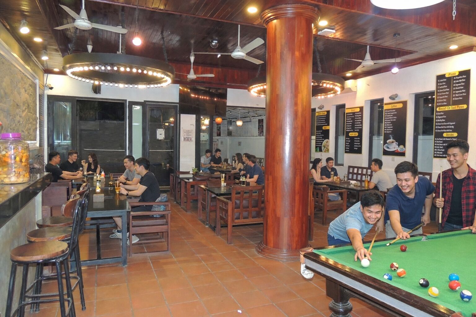 Una mesa de billar con jugadores, una zona de bar con taburetes y un tablero de menú en un restaurante.
