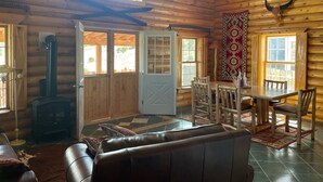 A log cabin interior with a fireplace, wooden dining table, and a large window.