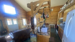 A log cabin interior with a wooden dining table, a sofa, a kitchen area with a refrigerator, and a hanging pot rack.