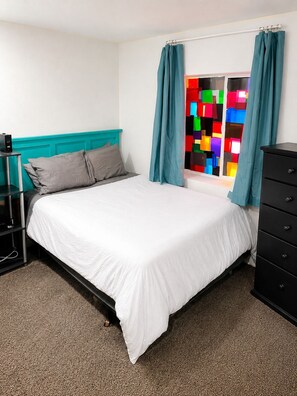 A bed with a white sheet, a teal headboard, grey pillows, and a black dresser.