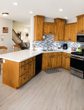 A kitchen with wooden cabinets, a white countertop, a stainless steel stove, and a tiled backsplash. - The Good Life (Cedar City)