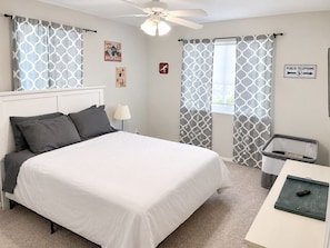A bedroom with a bed, a nightstand, a lamp, a ceiling fan, and a trash bin. - The Good Life (Cedar City)