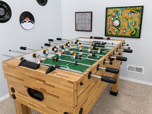 A wooden foosball table with a green playing surface, surrounded by wall decorations including a framed crossword puzzle and a colorful board game. - The Good Life (Cedar City)