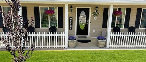 A two-story house with a white picket fence, a front porch with chairs, and a welcome sign.